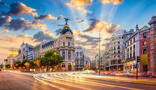 Gran Via in Madrid at sunset with historic architecture, illuminated street traffic, and dramatic golden clouds in blue sky